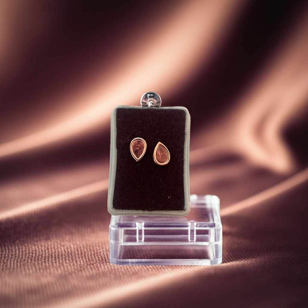 Primer plano de aretes broqueles de plata 925 y gota rosa, contrastando con el fondo oscuro y la base negra de exhibición.